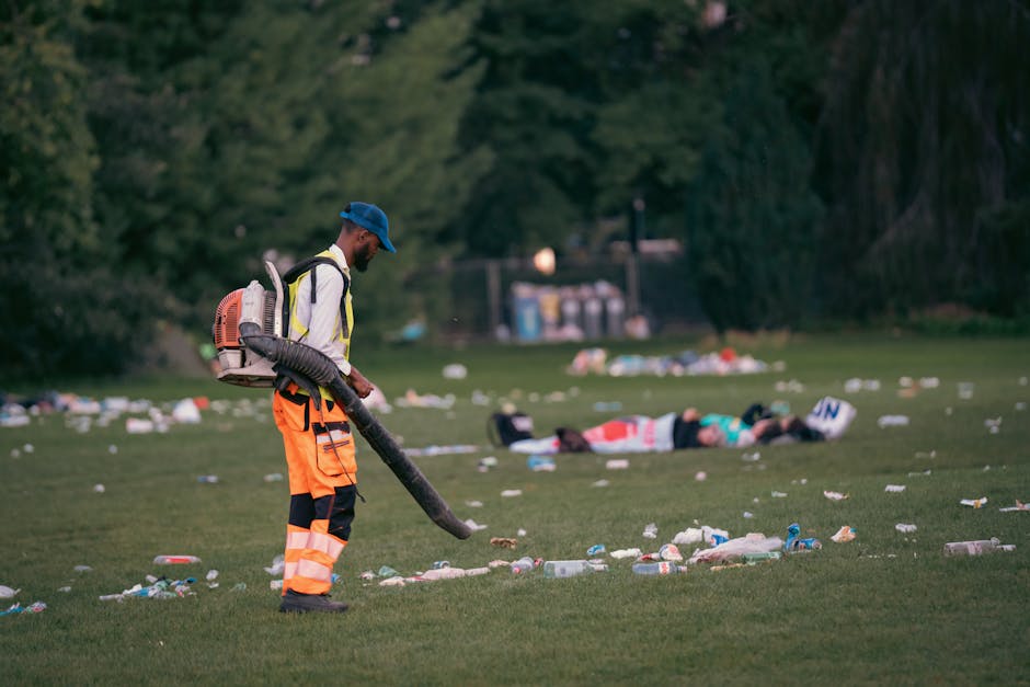 A worker dressed in high-visibility orange trousers with reflective strips, a light-colored jacket, and a blue cap is using a leaf blower to clear litter from a grassy park area. The worker is carryin
