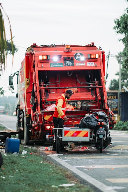 A worker dressed in high-visibility orange trousers with reflective strips, a light-colored jacket, and a blue cap is using a leaf blower to clear litter from a grassy park area. The worker is carryin